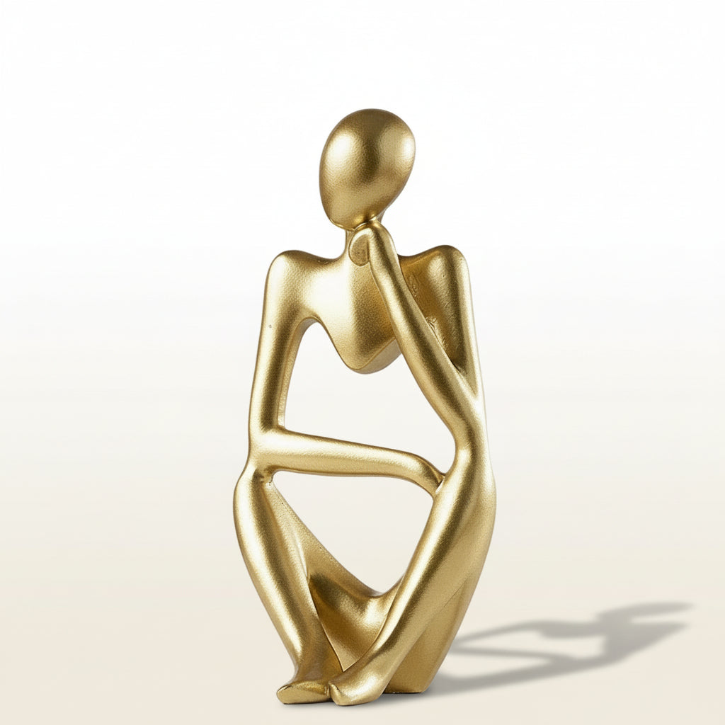 Gold abstract sculpture of a sitting figure on a wooden surface with a white background